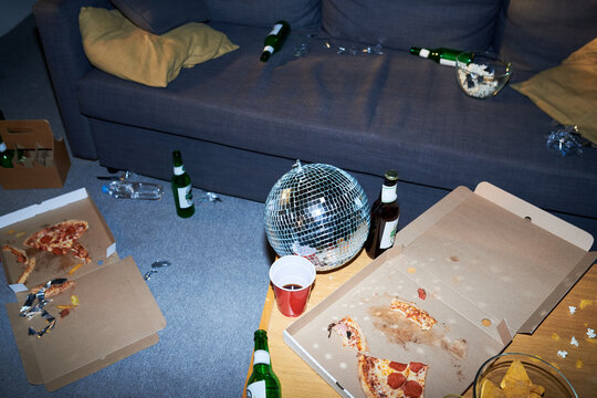 Messy living room after party showing empty pizza boxes, beer bottles, and disco ball on table. Concept of hangover or cleanup service, suitable for domestic cleaning ads, high angle view