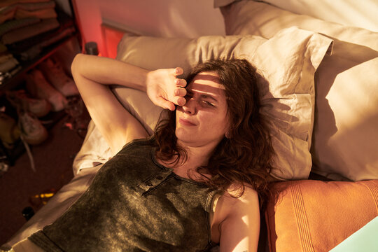 Caucasian young adult woman lying in bed shielding eyes from morning sunlight, waking up with difficulty. Concept of sleep deprivation, insomnia, and morning routine, high angle shot