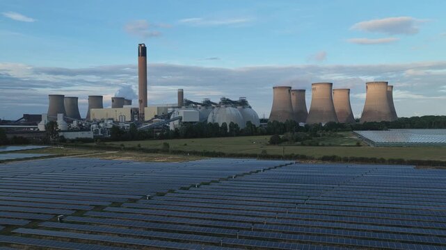 Drax Power Station in North Yorkshire features cooling towers and smoking chimneys rising above large solar panel fields under a clear blue sky during the bright late afternoon light.