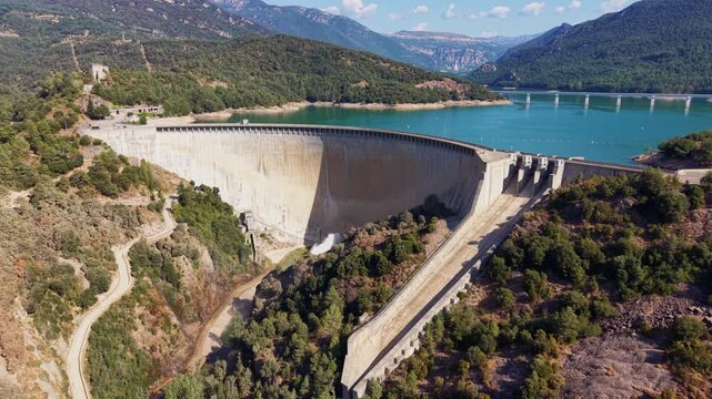 Baells dam in Catalonia, Spain, with a large concrete arch wall holding back a turquoise reservoir surrounded by green forested mountains under clear blue sky