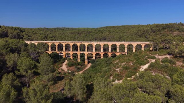 Ferreres Aqueduct in Tarragona, Spain, an ancient Roman aqueduct with double-tiered stone arches spanning a forested valley under a clear blue sky