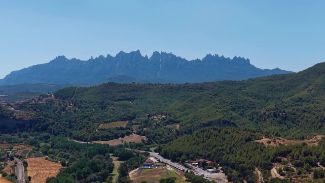 Montserrat mountain range with jagged rocky peaks rising above green forested hills, farmland, and a highway in Catalonia, Spain under clear blue sky