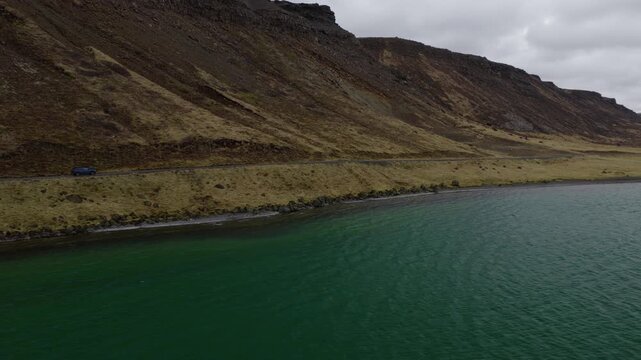 Car travels along Road 42 beside Kleifarvatn on the Reykjanes Peninsula, with volcanic slopes rising above the lakeshore in Grindavikurbaer, Iceland, drone tracking shot.