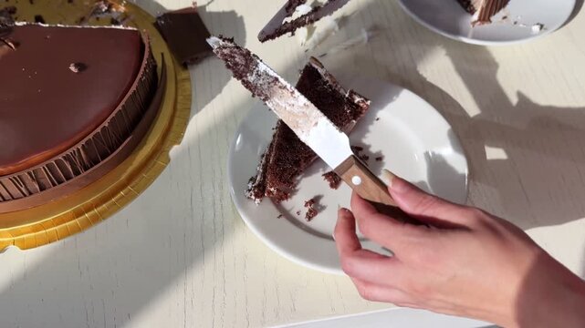 Close-up high angle shot of serving a slice of rich chocolate birthday cake with a knife onto a white plate. Partially eaten dessert rests on a golden tray on a white table in bright indoor sunlight.