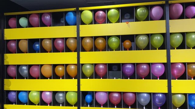 A vibrant array of colorful balloons arranged in rows on a bright yellow carnival balloon popper game wall. Shot indoors under artificial lighting, creating a fun, static recreational background.