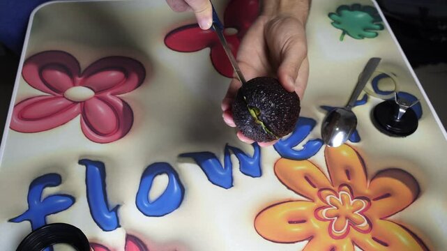 Top down close up of male hands holding two halves of a fresh green avocado over a colorful floral kitchen table. Preparing healthy vegetarian food indoors with a metal spoon visible nearby.