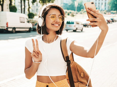 Fashion portrait of young stylish hipster woman walking in the street.  making selfie.Smiling model enjoy her weekends with backpack. Female listening to music via headphones