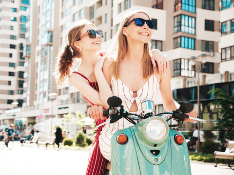 Two young beautiful smiling hipster female in trendy overalls.Sexy carefree women driving retro motorbike on the street background.Positive models having fun, riding classic Italian scooter in eyewear