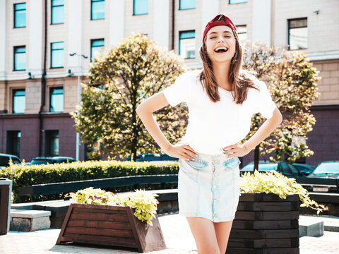 Young beautiful smiling hipster woman in trendy summer white t-shirt. Sexy carefree woman posing on the street background in cap at sunset. Positive model outdoors. Cheerful and happy