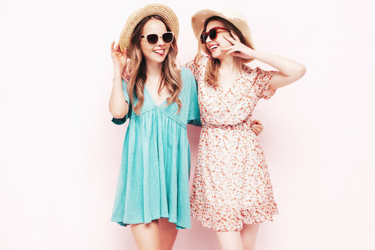 Two young beautiful smiling brunette hipster female in trendy summer dresses. Sexy carefree women posing near pink wall. Positive models having fun. Cheerful and happy. In hats and sunglasses