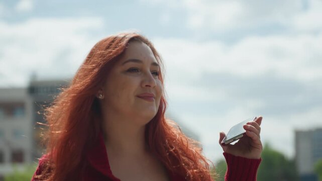 Close up of woman with long red hair wearing maroon knit sweater holding and looking at smartphone outdoors. Her expression is calm as woman interacts with device set against urban