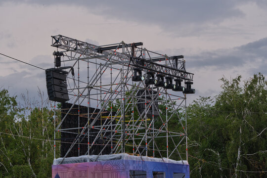 Outdoor festival stage metal framework with line array speakers, spot lights, and green trees behind.