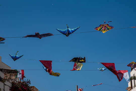 Kites and flags are stretched on lines across a blue sky at a street festival in San Vito lo Capo