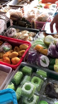 Slow motion vertical video of a woman's hand reaching to buy colorful traditional Indonesian sweet snacks known as jajanan pasar. Various delicious cakes are displayed in baskets at a local market.