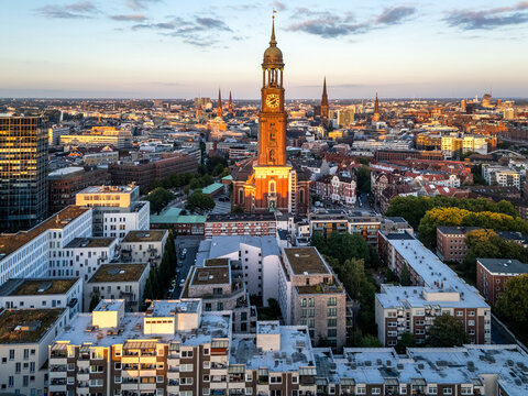 Aerial view of St. Michaeliskirche illuminated by golden hour light, surrounded by historic rooftops and the urban skyline of Hamburg, Germany.