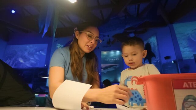 A young Latina mother and her 2-year-old son enjoy a creative moment, coloring sheets with sharks and fish designs in a blue LED-lit underwater-themed exhibit.