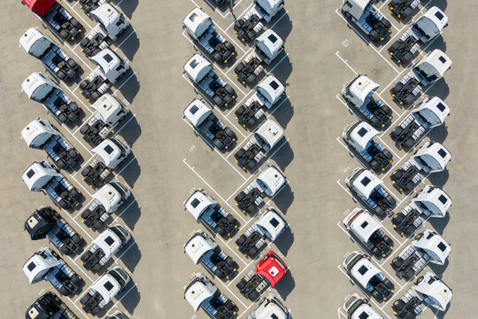 Aerial view of rows of parked semi-truck cabs with one red truck among many white ones in a paved lot in Poland.