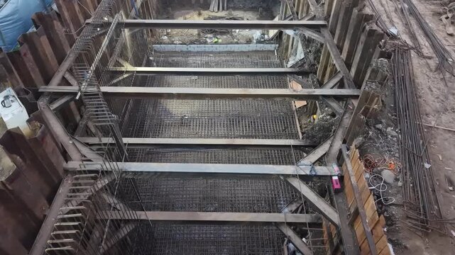 Workers build foundations near a calm river. Steel beams and wooden frames stand tall. pipes lie scattered on dirt ground. People move carefully through the construction site street construction