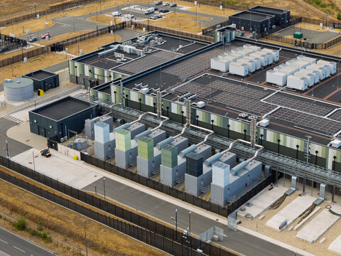 Aerial view of a modern data center facility featuring rows of cooling units and solar panels on the roof, surrounded by a secure fenced perimeter in the United Kingdom.