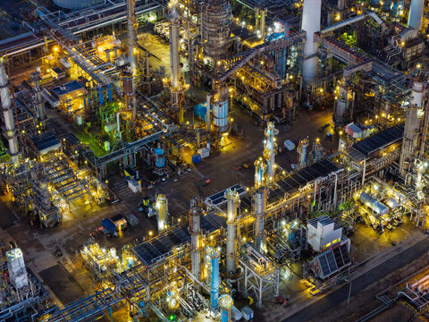 Aerial view of an industrial oil refinery at night, featuring illuminated distillation towers, complex pipe networks, storage tanks, and metal structures in the United Kingdom.