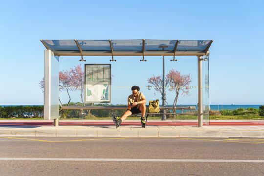 Latino man in rollerblades resting at a bus stop on a coastal road, checking phone