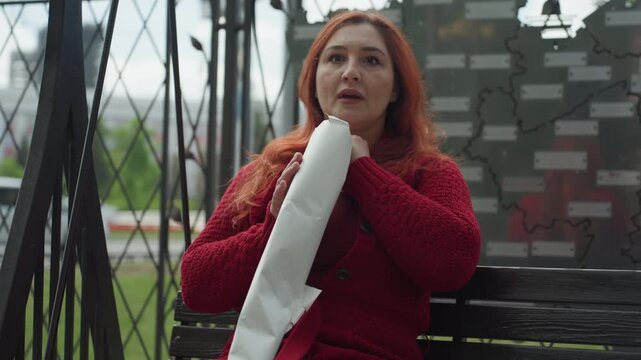 Close up of woman with long red hair wearing textured knit sweater holding and examining unrolled white paper scroll while seated on wooden bench outdoors. background features blurred