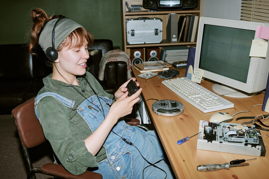 Young adult Caucasian woman wearing headphones and holding old pager in retro office, repairing vintage computer hardware. Image suitable for nostalgic tech marketing campaigns