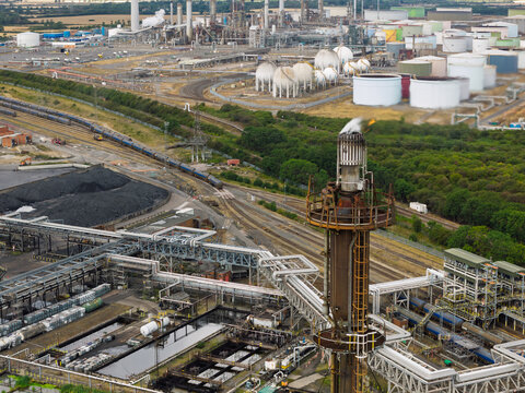 Aerial view of an industrial oil refinery with a prominent flare stack, large storage tanks, complex pipe networks, and railway tracks in the United Kingdom.