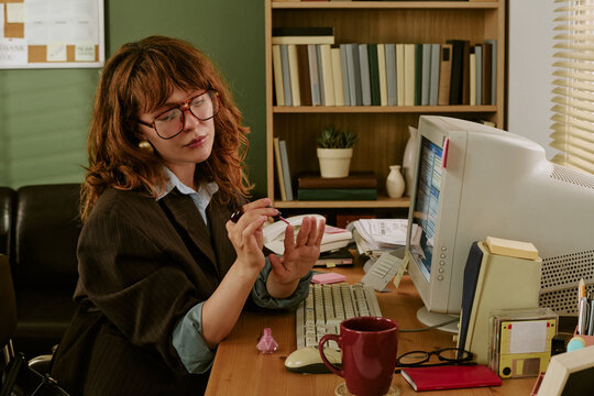 Young adult Caucasian woman applying nail polish at desk in retro office, slacking off during work hours. Image represents workplace distraction and is suitable for HR articles