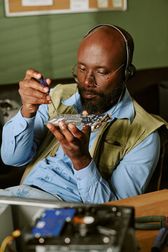 Vertical image of middle aged African American man wearing headphones and repairing computer circuit board in retro office, representing hardware engineering and technical support services