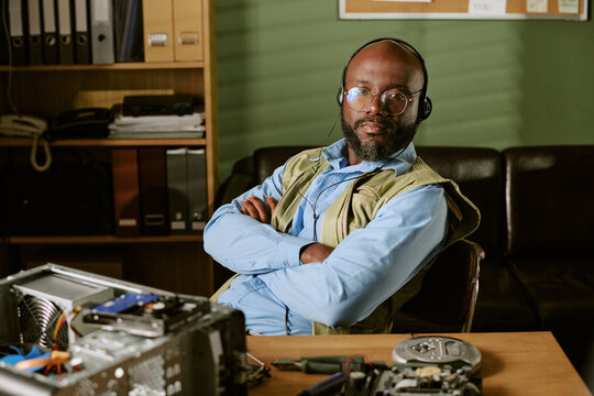 Portrait of middle aged African American man wearing headset and sitting with arms crossed in retro office. Image represents tech support and is suitable for tech service marketing