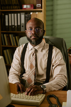 Portrait of middle aged African American man working on retro computer in vintage office. Image represents corporate history and is suitable for business heritage campaigns, vertical