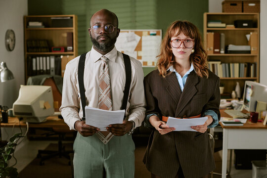 Portrait of African American middle aged man and Caucasian young woman standing in retro office holding documents. Image represents vintage corporate culture and diverse teamwork
