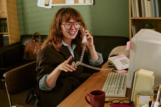 Young adult Caucasian woman working in retro office, talking on vintage telephone and smiling. Image suitable for nostalgic corporate communication and vintage business marketing