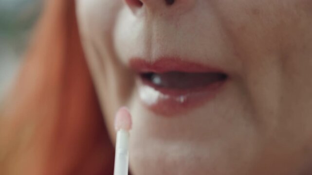 Close up of woman applying lip gloss to her lips with steady hand and applicator. Lips are shown in detail as product is spread evenly across them highlighting texture change from dry