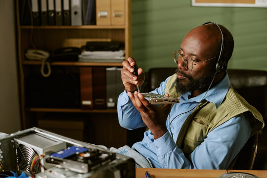 Middle aged Black man wearing headset repairing computer hardware in retro office. Image represents technical support and tech maintenance services, suitable for corporate marketing