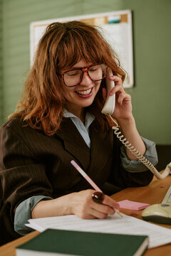 Young adult Caucasian woman working in retro office, talking on landline telephone and writing notes. Image suitable for vintage corporate communication campaigns, vertical