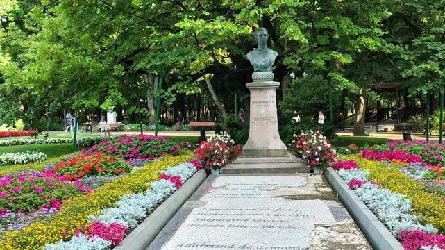 Bust of Mihai Eminescu monument with poetry mosaic paved path surrounded by colorful flower beds in a sunlit public park.