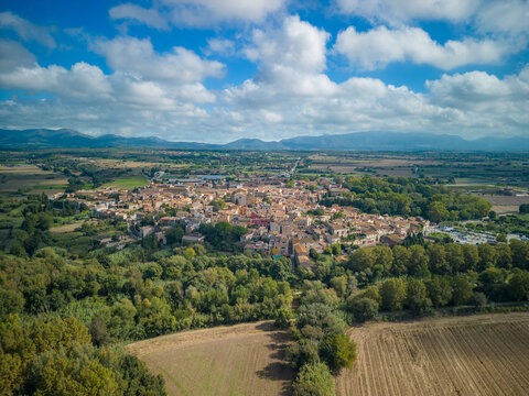 Aerial view of the historic town of Peralada surrounded by lush green trees, agricultural fields, and distant mountains under a blue sky in Peralada, Catalonia, Spain.