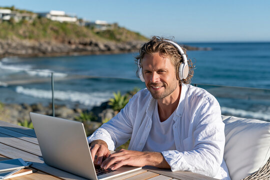 Man smiles while working on laptop by ocean. White headphones frame his relaxed, content expression. Sunny coastal view behind him, with calm waves and distant cliffs. Perfect for remote work