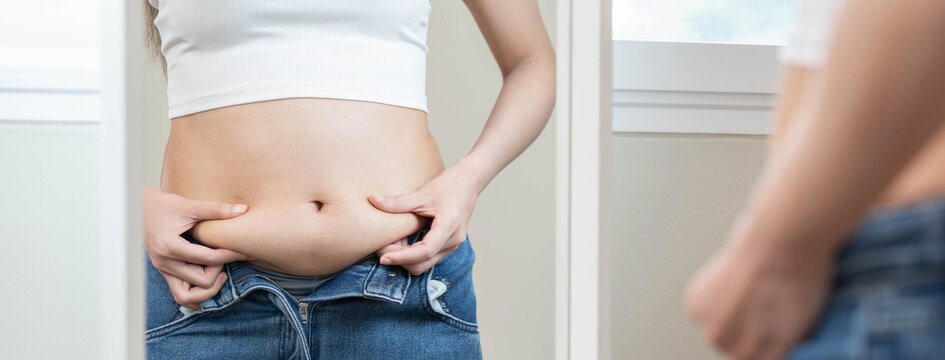 Overweight female trying to button up tight denim trousers after weight gain.