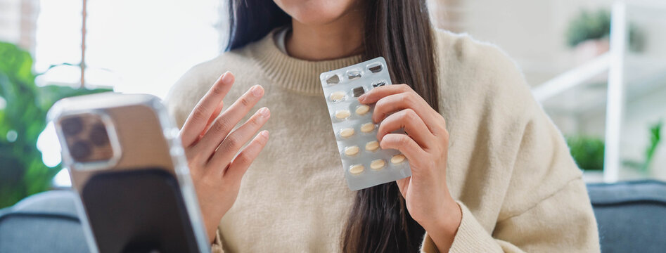 Sick Asian woman holding medicine pill blister pack during online video call telehealth consultation with doctor via smartphone on sofa at home