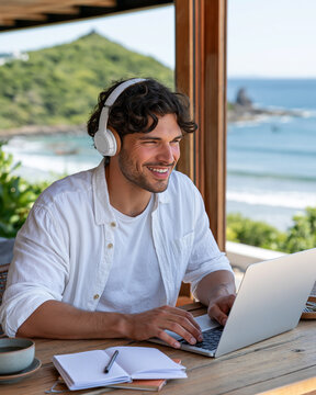 Man smiles, wearing headphones, focused on laptop. Laptop sits on wooden table, beside notebook and pen. Ocean waves roll behind him, green hill rises in distance. Natural light bathes scene