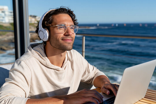 Man relaxes with headphones and laptop by the sea. He smiles softly, enjoying calm, sunny weather. Ocean waves roll gently behind him. Laptop rests on wooden table near window. Perfect for remote work