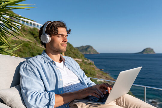Man relaxes on balcony, using laptop, wearing headphones. Blue sky, ocean, and distant islands provide scenic backdrop. He appears calm, focused, enjoying quiet time outdoors. Perfect for remote work