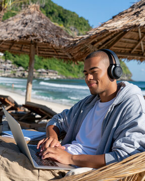Man relaxes on beach, typing on laptop. He wears headphones, smiling gently. Tropical scene, waves crash behind him. Sheltered by thatched umbrellas, he enjoys solitude