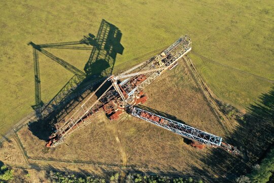 Aerial view of a massive, rusty bucket-wheel excavator situated in a field in the Lausitz region, casting a long shadow across the landscape in Brandenburg, Germany.
