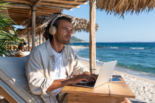Man works on laptop at beachside table. He wears headphones, focused on screen. Sunlight glints off calm ocean waves. Thatched roof shelters him from sun. Tropical vibe with gentle sea breeze