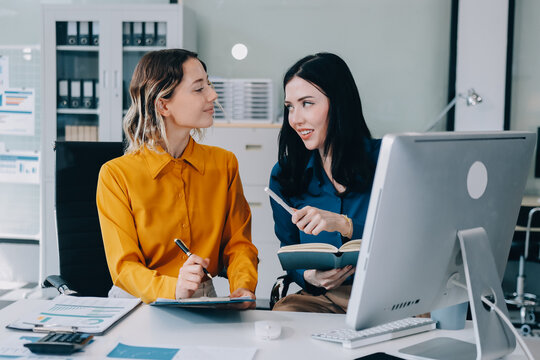 Two businesswomen discussing work using a laptop in a modern office lounge area, showing teamwork and communication