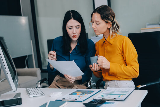 Two businesswomen discussing work using a laptop in a modern office lounge area, showing teamwork and communication